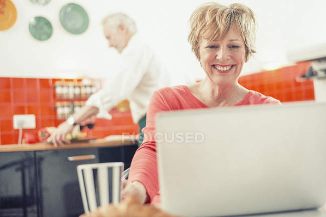 Зрелая женщина использует ноутбук на кухне с мужчиной на заднем плане — стоковое фото