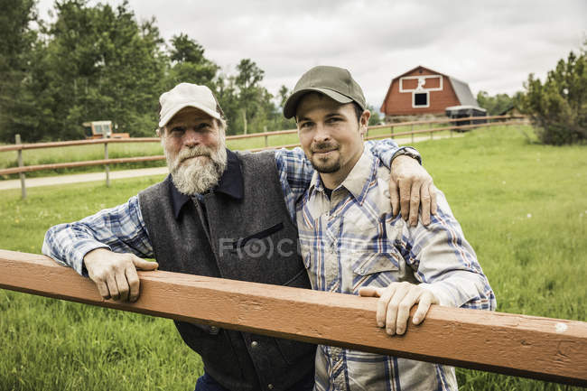 Мужчина на ферме, прислонившись к забору, обнимает сына и смотрит в камеру улыбаясь. — стоковое фото