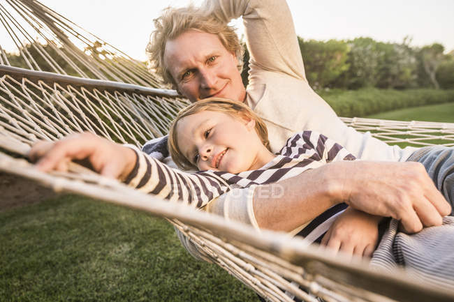 Padre e hijo en el jardín tumbados en hamaca mirando a la cámara abrazando y sonriendo - foto de stock