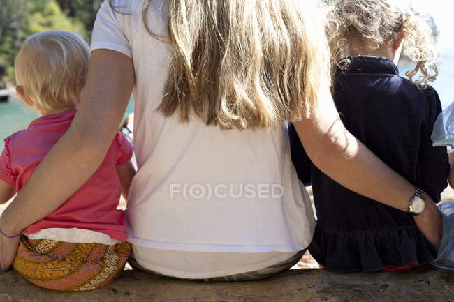Vista trasera de la madre y dos hijas sentadas en el tronco del árbol en la playa, Nueva Zelanda - foto de stock