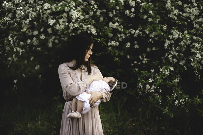 Mid adult woman carrying baby daughter in arms by garden apple blossom — Stock Photo