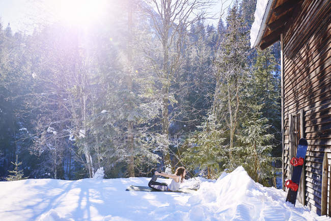 Woman practicing bow yoga pose by log cabin in snow,  Austria — Stock Photo