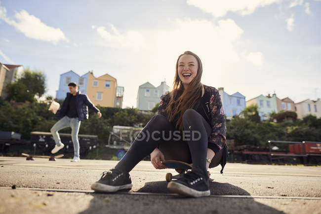 Dos amigos jugando al aire libre, mujer joven sentada en el monopatín, riendo, Bristol, Reino Unido - foto de stock