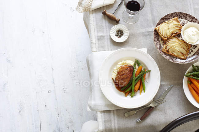 Top view of table meal with plates of duck breast, carrots, asparagus and mashed potato, and apple tart — Stock Photo