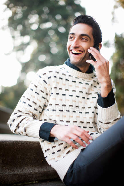 Молодий чоловік сидить і використовує мобільний телефон, посміхаючись — стокове фото