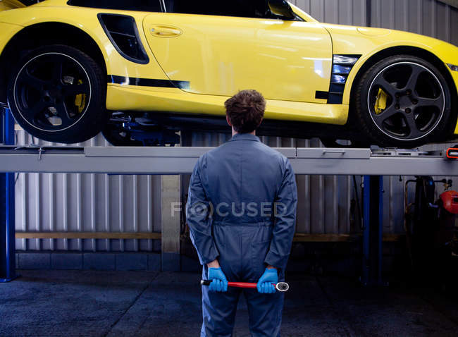 Mécanicien mâle vérifiant voiture de sport jaune, tenant clé à douille derrière son dos, vue arrière — Photo de stock