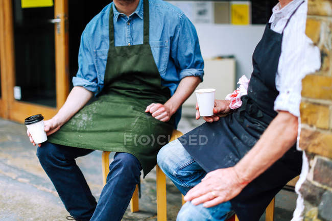 Старший ремесленник пьет кофе и болтает с молодым человеком за пределами мастерской, в середине секции — стоковое фото