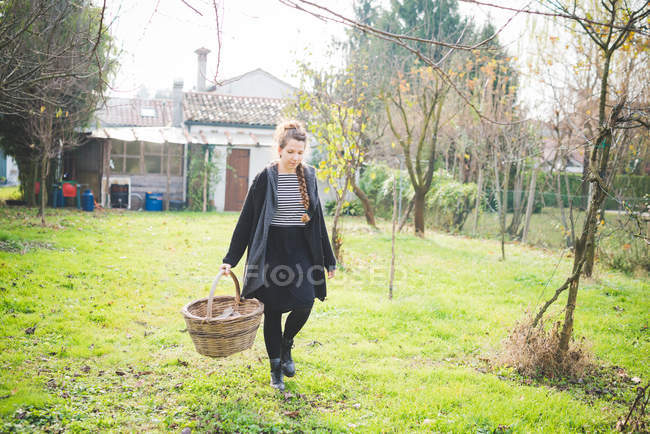Visão frontal de comprimento total da jovem mulher no jardim carregando cesta wickerwork — Fotografia de Stock
