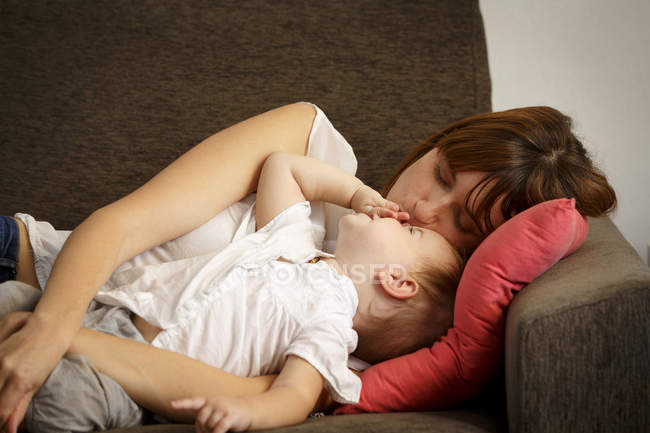 Mid adult woman reclining and kissing toddler daughter on sitting room sofa — Stock Photo