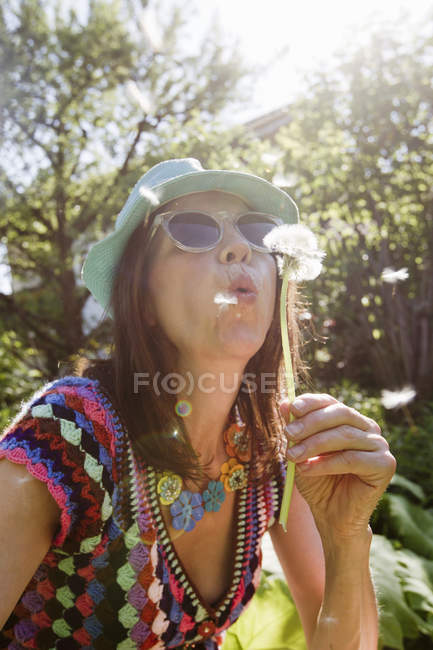 Mature woman wearing sunhat and sunglasses blowing dandelion seeds — Stock Photo