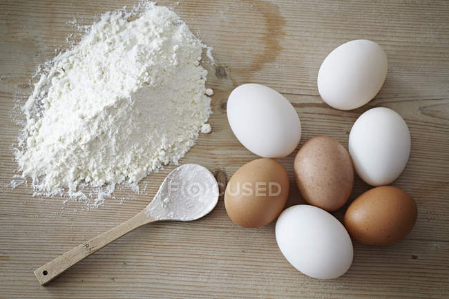 Ovos e farinha com colher de madeira na superfície de madeira — Fotografia de Stock