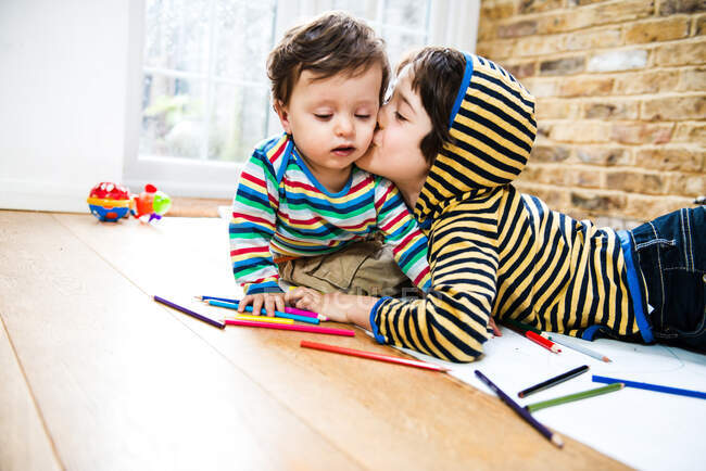 Boy kissing baby brother while lying on floor drawing — Stock Photo
