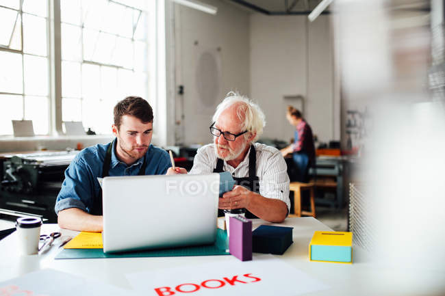 Ein älterer Handwerker und ein junger Mann schauen in der Buchkunstwerkstatt auf den Laptop — Stockfoto