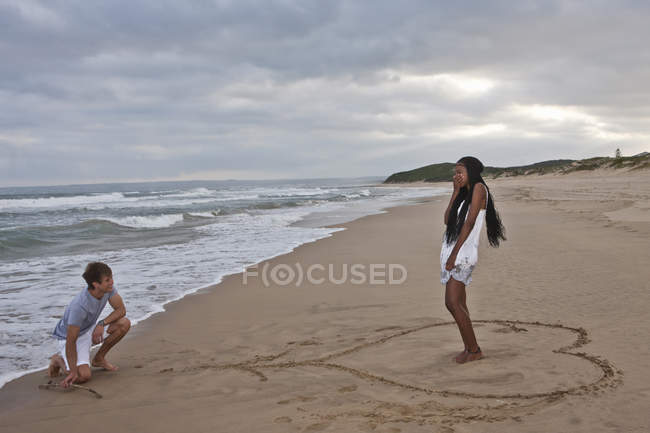 Молодой человек на коленях на пляже, молодая женщина, стоящая в форме сердца — стоковое фото