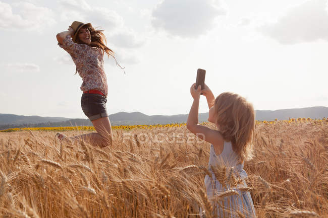 Girl taking photograph of mother in wheat field jumping — Stock Photo