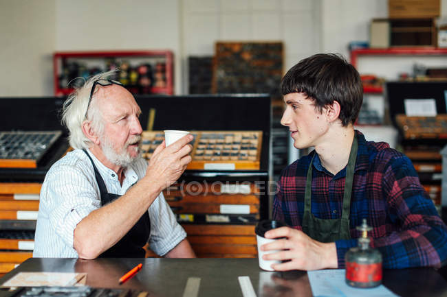 Senior craftsman drinking coffee and chatting to young craftsman in book arts workshop — Stock Photo