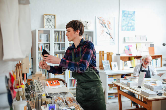 Молодий майстер збирає пензлик і дивиться на матеріали в крамниці, з старшим чоловіком на задньому плані — стокове фото