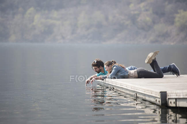 Young couple lying on pier dipping fingers in Lake Mergozzo, Verbania, Piemonte, Italy — Stock Photo