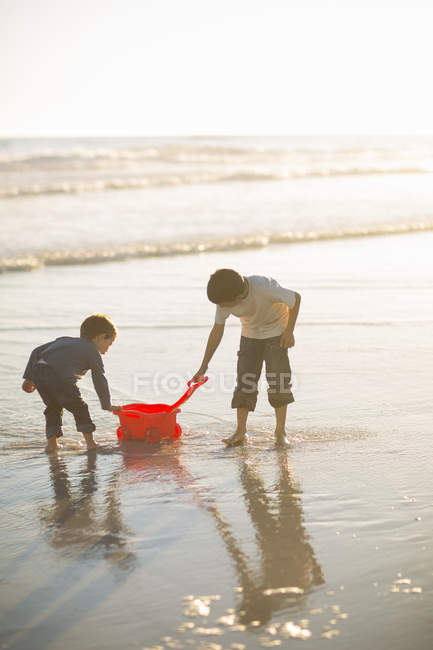 Irmãos enchendo caminhão de brinquedo com água do mar na praia — Fotografia de Stock
