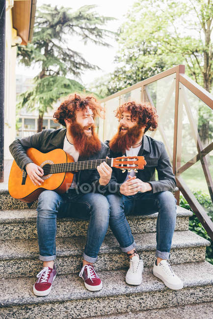 Young male hipster twins with red beards sitting on stairway playing guitar — Stock Photo