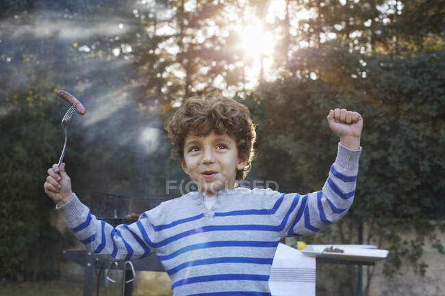 Menino braços levantados segurando salsicha no garfo sorrindo — Fotografia de Stock