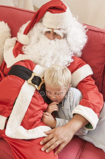Homme assis sur canapé habillé comme Père Noël étreignant garçon — Photo de stock