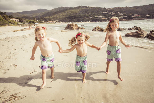 Три молодих сестри тримаються за руки, бігають уздовж пляжу, посміхаючись — стокове фото