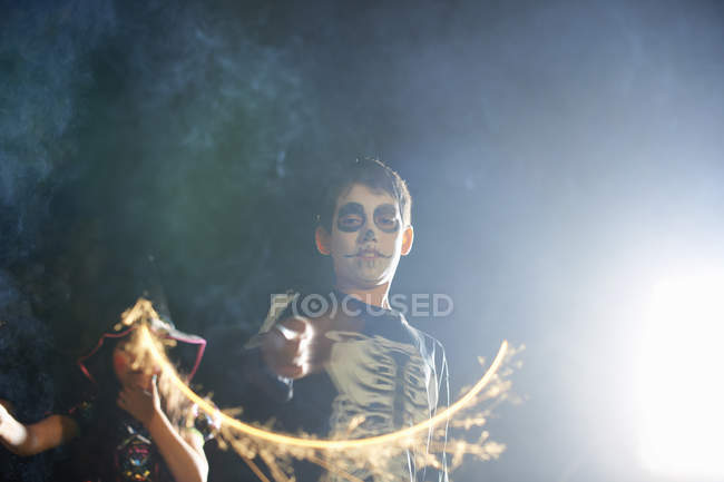Брат и сестра в костюмах на Хэллоуин играют с бенгальскими огнями в саду ночью — стоковое фото