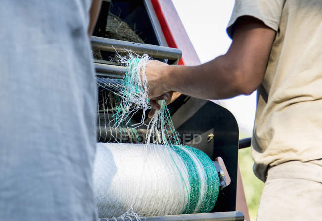 Fotografía recortada de dos trabajadores agrícolas masculinos desenredando redes en empacadora de heno - foto de stock