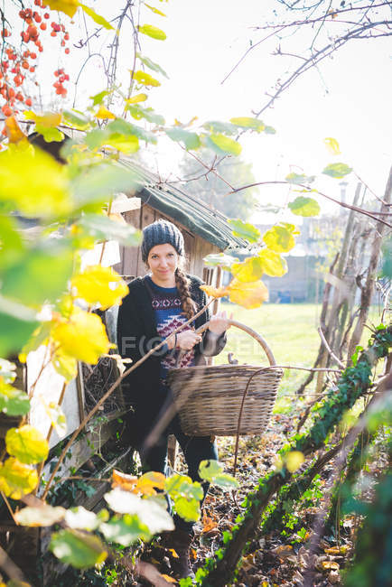 Vue à travers les feuilles de la jeune femme dans le jardin portant un chapeau en tricot tenant panier en vannerie regardant la caméra sourire — Photo de stock