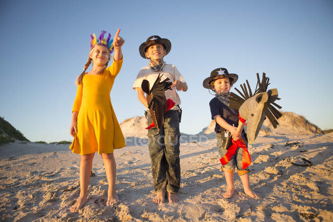 Hermana y hermanos vestidos como nativos americanos y vaqueros señalando desde dunas de arena - foto de stock