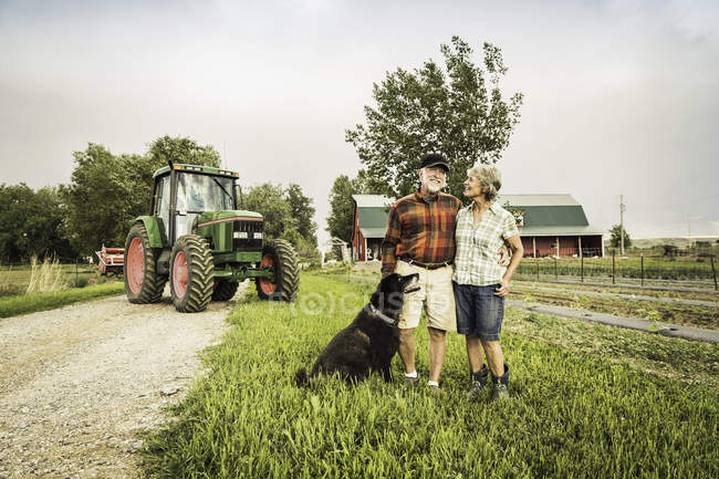 Ehepaar mit Hund auf Bauernhof vor Traktor und blickt lächelnd in Kamera — Stockfoto
