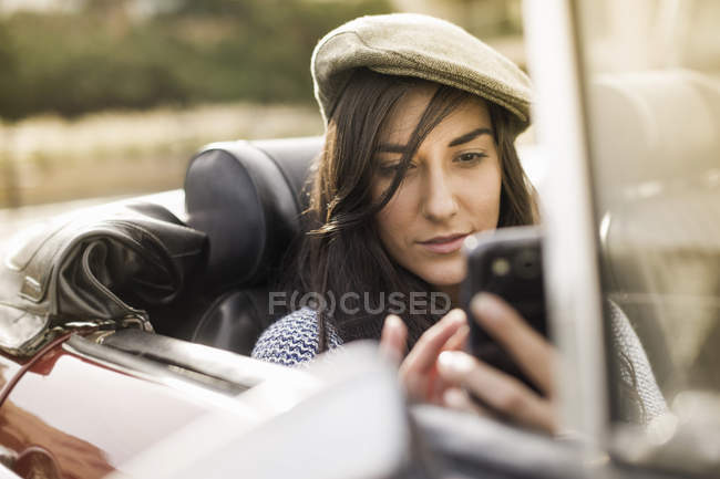 Молода жінка носить плоску шапку в кабріолеті за допомогою смартфона — стокове фото