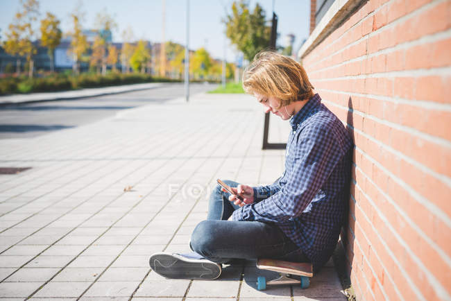Молодий чоловік міський скейтбордист сидить на тротуарі вибираючи музику смартфона — стокове фото