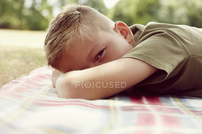 Vue de niveau de surface du garçon allongé sur la couverture de pique-nique, visage obscurci en regardant la caméra — Photo de stock
