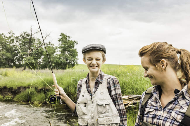 Madre e hija por río sosteniendo cañas de pescar sonriendo - foto de stock
