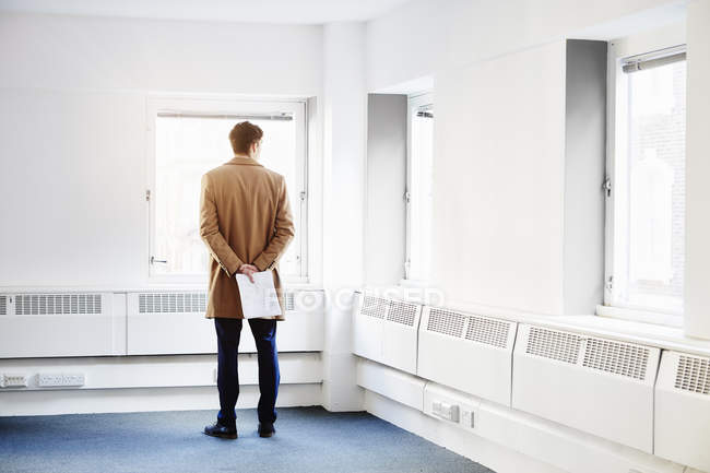 Rear view of man in empty office, hands behind back looking out of window — Stock Photo