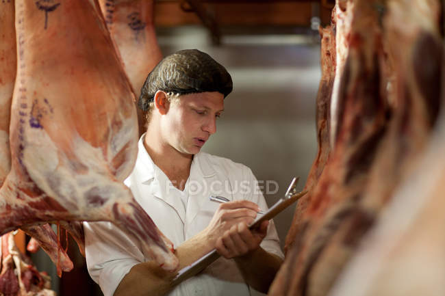 Carnicero macho con portapapeles inspeccionando carne - foto de stock