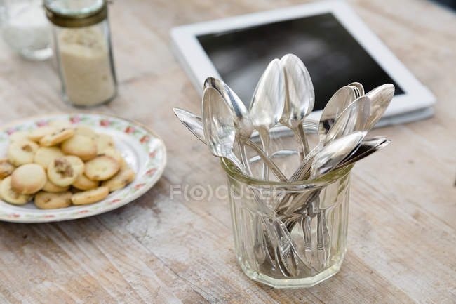 Tableta digital, plato de galletas y tarro de cucharas sobre superficie de madera - foto de stock