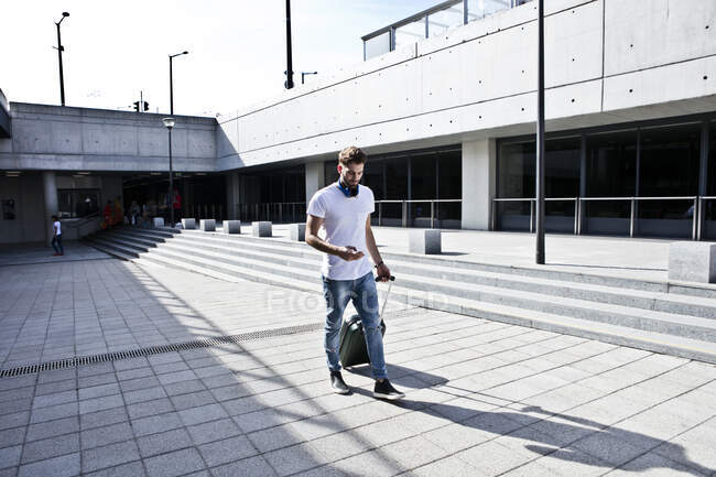 Молодой человек выходит на улицу, вытаскивает чемодан с колёсами, используя смартфон — стоковое фото