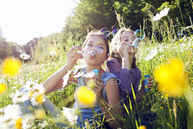 Sisters sitting in field of flower blowing bubbles — Stock Photo