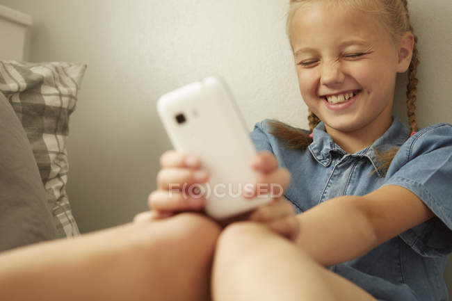 Дівчина сидить, спираючись на стіну тримає смартфон, очі закриті посміхаючись — стокове фото