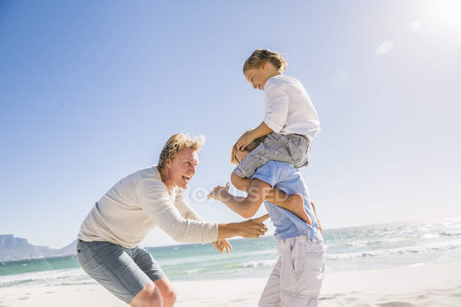 Big brother on beach carrying brother on shoulders, father tickling — Stock Photo
