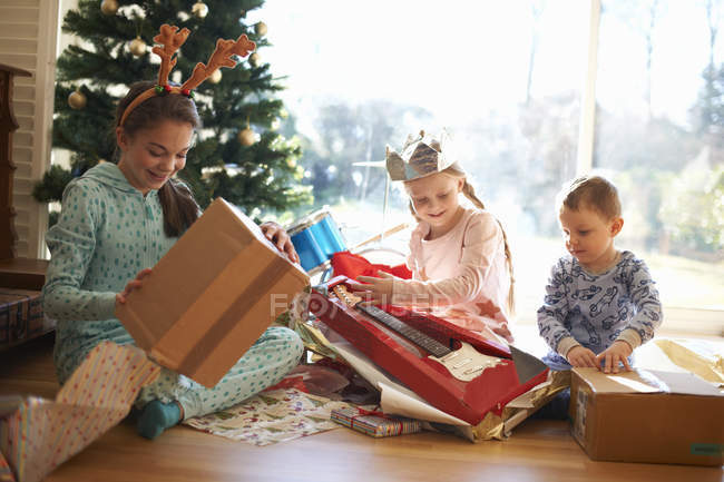 Menino e irmãs sentados na sala de estar chão abrindo presentes de Natal — Fotografia de Stock