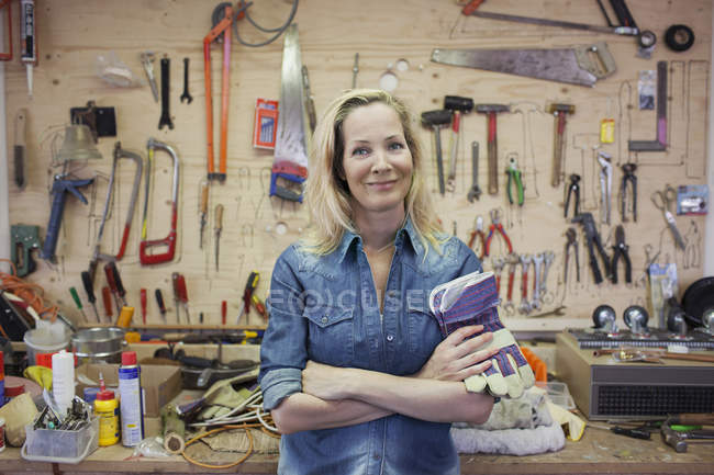Mujer en el taller, brazos cruzados sosteniendo guantes protectores mirando a la cámara sonriendo - foto de stock
