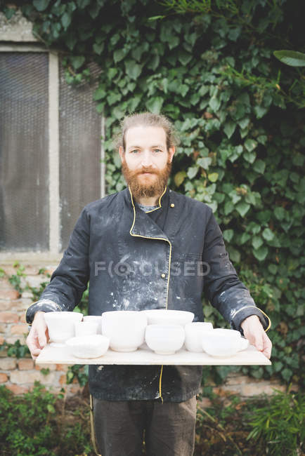Front view of bearded mid adult man holding tray with clay pot looking at camera smiling — Stock Photo