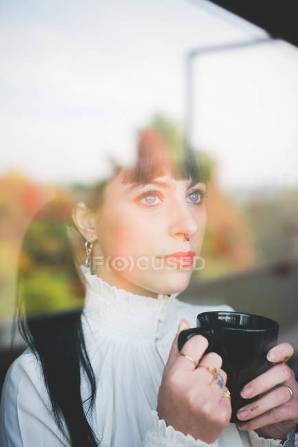Молода жінка з чашкою дивиться з вікна — стокове фото