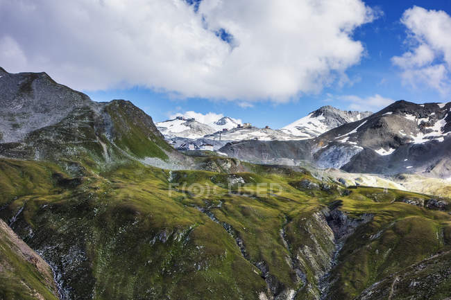 Green hills and snowcapped rocky mountains under cloudy sky — Stock Photo