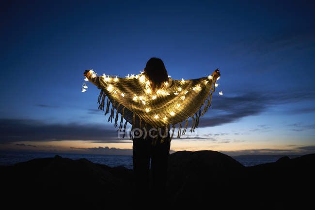 Задній вид жінка, загорнуті в шаль з зірка фея вогнями на пляжі вночі — стокове фото
