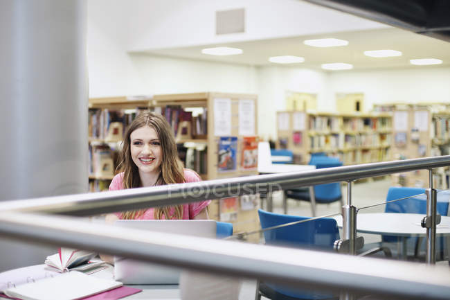 Mujer joven trabajando en la biblioteca, sonriendo y mirando hacia otro lado - foto de stock